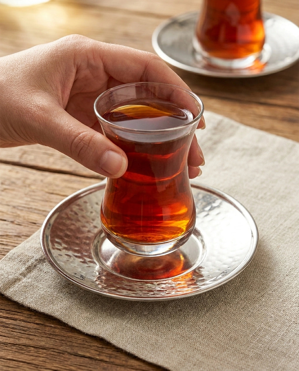 Gümüş renkli bakır altlık üzerinde duran çay dolu bardağı tutan bir el, arka planda ahşap masa. - Gaziantep Bakırcılık