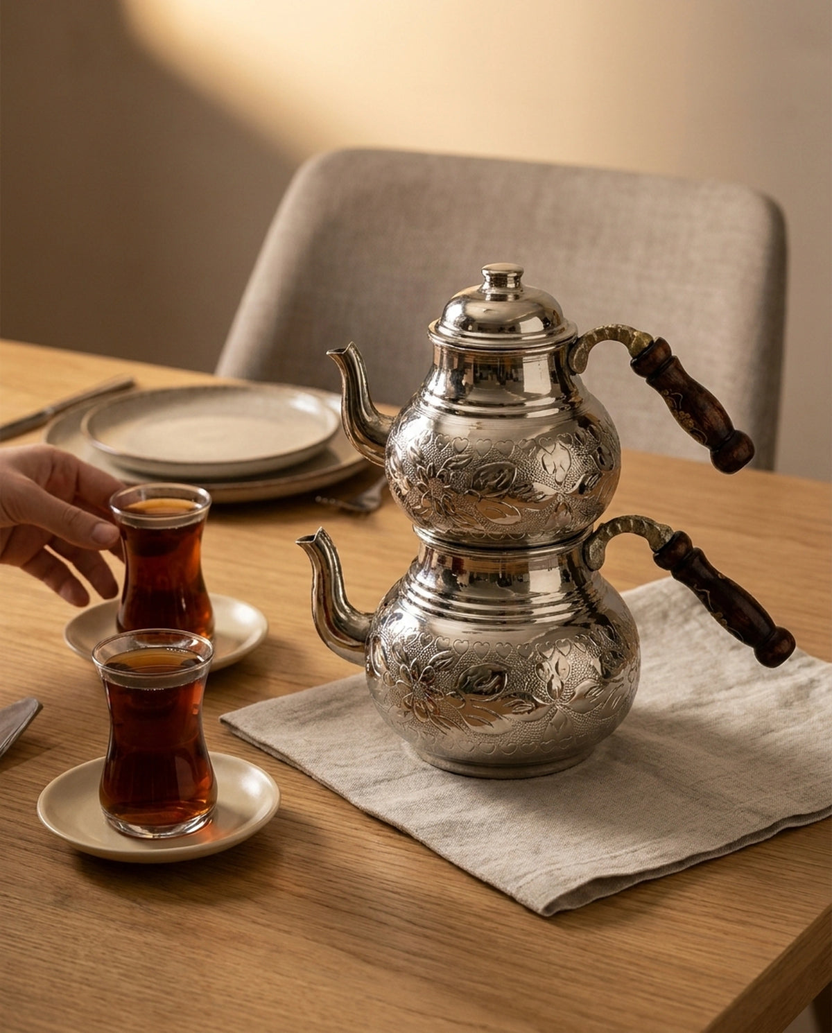 Ahşap masada duran işlemeli çaydanlık ile uzanan bir el ve iki bardak çayın görünümü. - Gaziantep Bakırcılık