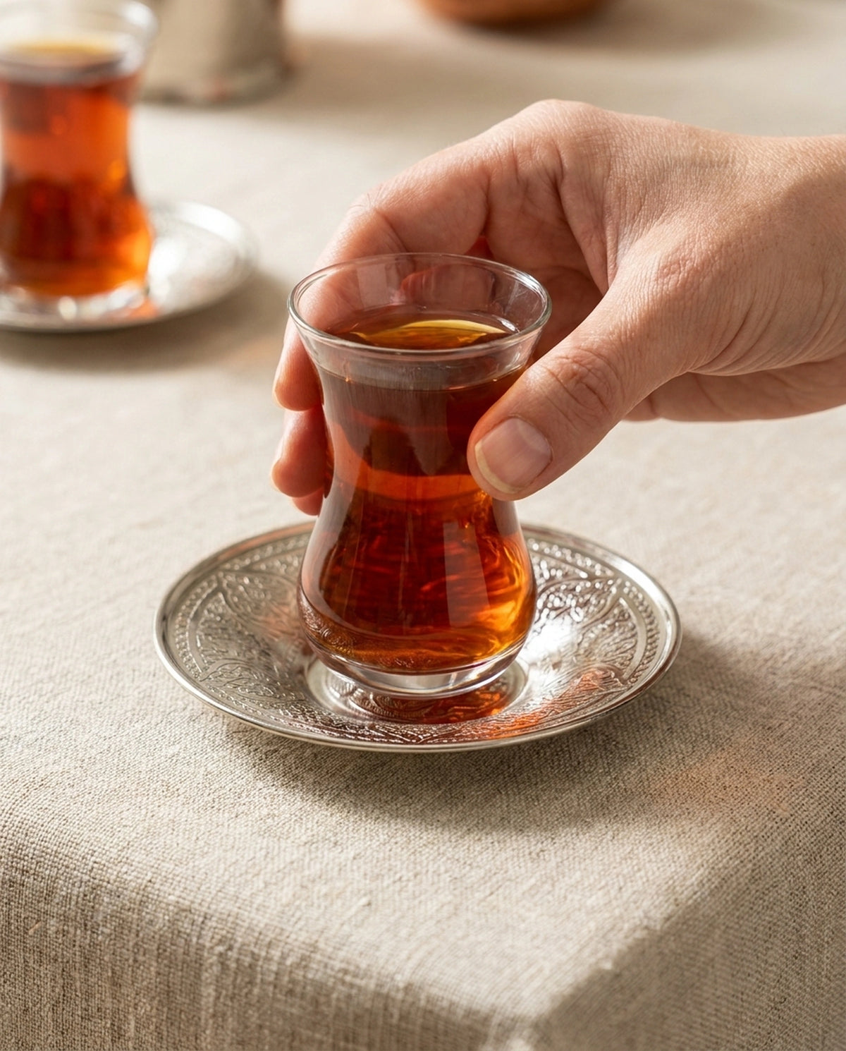 Keten örtülü masada işlemeli gümüş renkli bakır çay tabağına çay bardağını yerleştiren el. - Gaziantep Bakırcılık