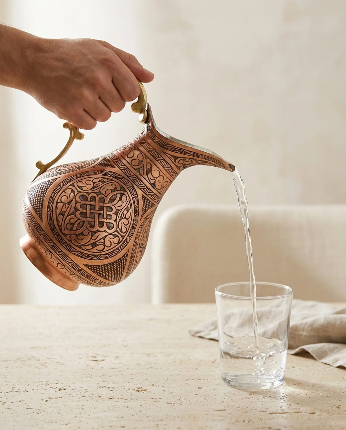 Bir elin pirinç kulptan tutarak cam bardağa su doldurduğu işlemeli bakır sürahi kullanım anı. - Gaziantep Bakırcılık