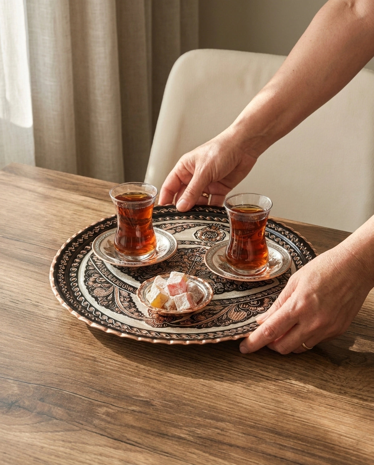 İki çay bardağı ve lokumluk taşıyan siyah bakır tepsinin ellerle ahşap masaya servis edilişi. - Gaziantep Bakırcılık