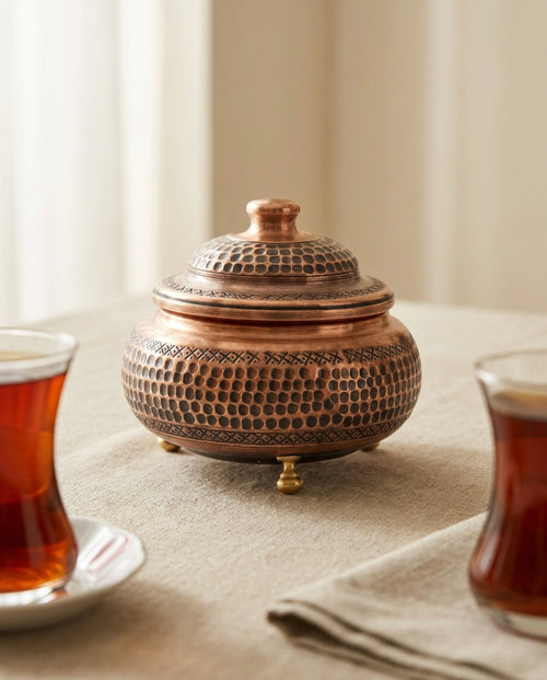 Masa örtüsü üzerinde iki bardak çay arasında sergilenen yuvarlak formlu eskitme bakır şekerlik. - Gaziantep Bakırcılık