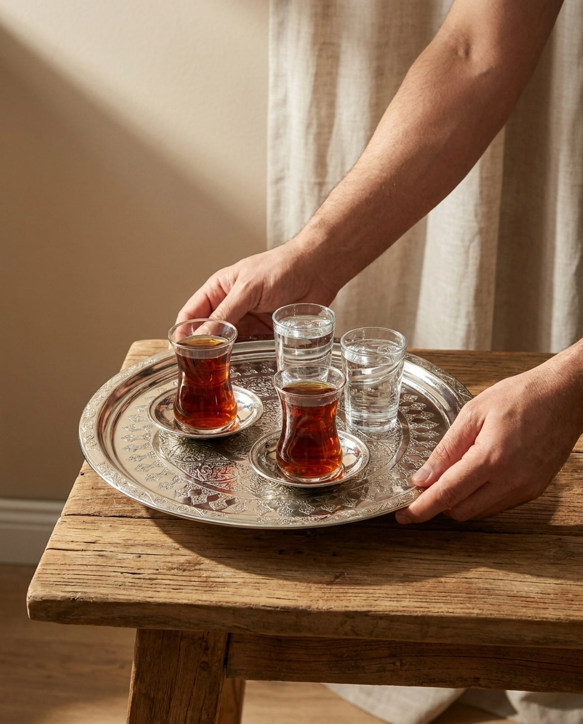 Ahşap masa üzerinde iki bardak çay ve su taşıyan gümüş renkli tepsiyi kenarlarından tutan eller. - Gaziantep Bakırcılık