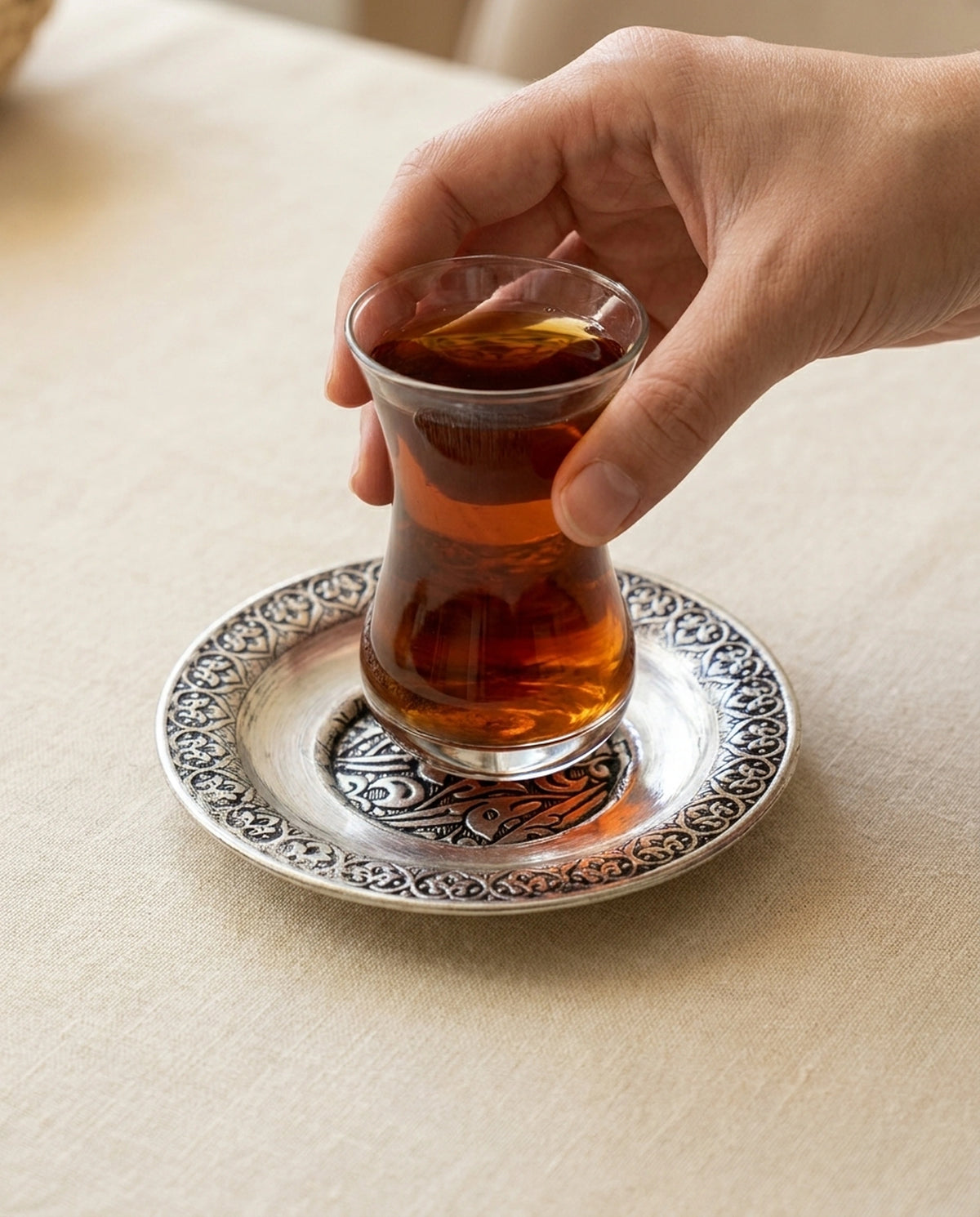 Bir elin işlemeli gümüş renk bakır çay bardak altlığı üzerindeki dolu çay bardağını tutuş detayı. - Gaziantep Bakırcılık