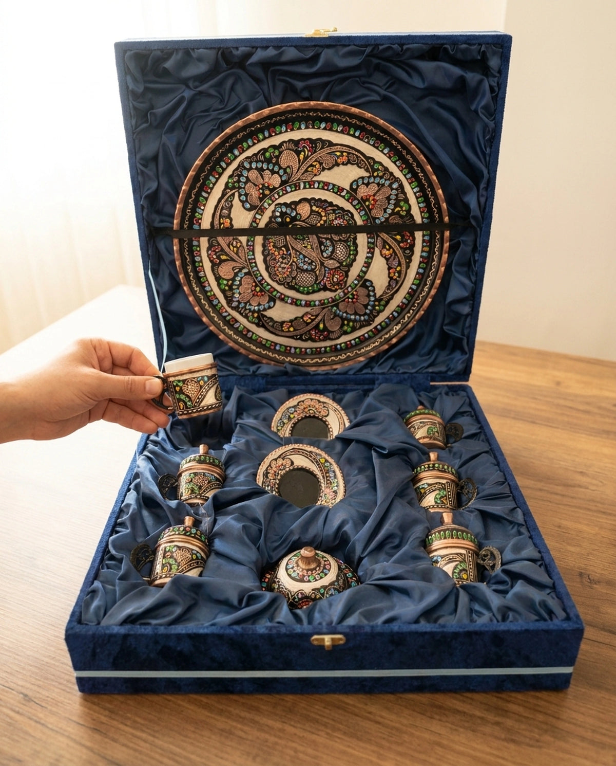 Bir elin rumi motifli bakır kahve fincanını porselen haznesiyle birlikte mavi kadife kutudan alırken görünümü. - Gaziantep Bakırcılık